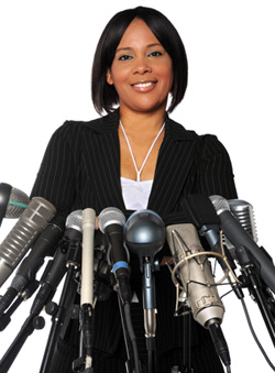 A woman is standing in front of a podium at a press conference. There are a number of microphones in front of her.