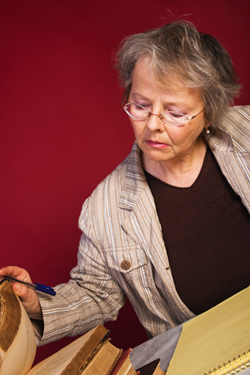 A female writer is skimming through some novels at her desk. In front of her is a notepad and she is holding a pen. She is reading about how to improve her fiction writing. 