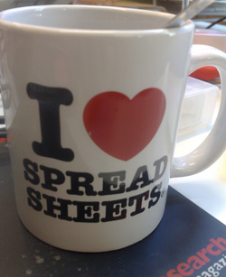 Our editors explain how to track changes in Excel. A coffee mug is sitting on a table. Written on the mug is the saying "I Love Spread Sheets" where "love" is written as a heart. This shows how much the person using the mug loves track changes in Excel.