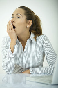 A woman is sitting at her desk. Her hand is in front of her mouth and she is stifling a yawn.