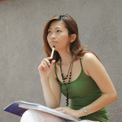 Our editors provide an example of an APA title page. A female student wearing a green tank top is sitting outside, with a notebook on her lap, contemplating how to write an APA title page.