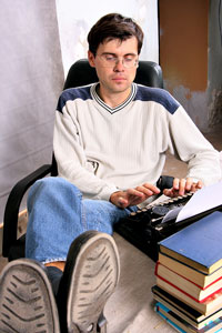 Our editors explain how to write a book proposal for fiction. A man leaning back in a chair and typing a book proposal; the table is covered with books.