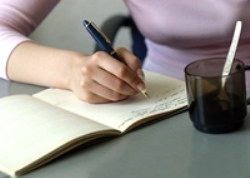Our editors provide tips to help you avoid telling writing. A woman is sitting at a desk with a cup of coffee (you cannot see her head) and is writing in a book.