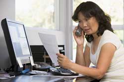 Our editors provide an example complaint. A woman is sitting at her computer holding an example complaint letter and talking to a customer service representative on the telephone.