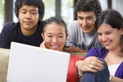 Four culturally diverse students sitting around a computer smiling. They are working on a homophone worksheet.