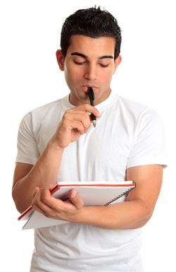 A young man in a white t-shirt is holding a black pen up to his lips with one hand and a notebook in the other hand.