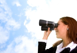 Our editors explain how to write a CV. A brunette woman in a black shirt is holding binoculars and looking out at a blue sky with white fluffy clouds in it.