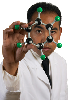 A student is standing in front of a white background. He holds a model of an organic compound in front of him. He is preparing to write his lab report or scientific paper.