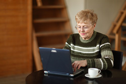 An older woman is sitting at a table with her laptop and a cup of coffee. She is writing a novel for National Novel Writing Month.