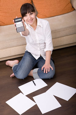 Our editors offer helpful tax tips for writers. A young brunette woman is sitting on the floor of her house holding a calculator; there are pieces of paper that contain tax tips on the floor.